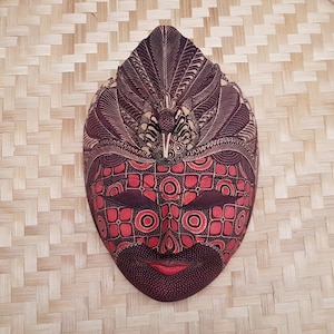 Pode incluir: Uma máscara tradicional indonésia com padrões intrincados em vermelho, preto e branco. A máscara apresenta um rosto estilizado com os olhos fechados e uma pequena boca.