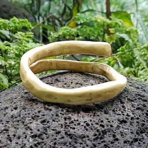 Peut inclure: Un bracelet clair fait à la main avec un design ouvert et unique. Le bracelet est fait d'un matériau lisse et organique et repose sur une pierre sombre et texturée. Le fond présente un feuillage vert luxuriant, créant une esthétique naturelle.