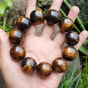 Puede incluir: Un brazalete hecho de cuentas de madera marrón oscuro con motas doradas. Las cuentas son redondas y tienen un acabado liso y pulido.