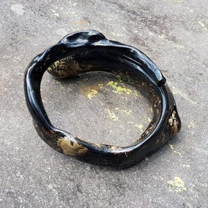 May include: A black, organic-shaped bracelet with a glossy finish. The bracelet has a unique, textured design with hints of gold and brown. The jewelry is set against a gray, textured surface.