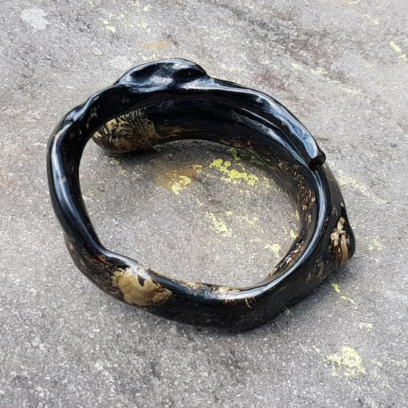 May include: A black, organic-shaped bracelet with a glossy finish. The bracelet has a unique, textured design with hints of gold and brown. The jewelry is set against a gray, textured surface.
