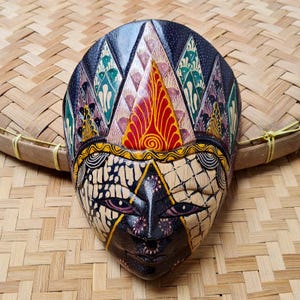 Può includere: Maschera di legno intagliata a mano con dettagli intricati. La maschera presenta una combinazione di colori nero, rosso, giallo e verde con motivi geometrici e floreali. È posta su uno sfondo di bambù intrecciato.