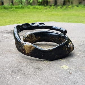 May include: A handcrafted, dark-stained wooden bracelet with a unique, organic shape. The bracelet features a swirling design with visible wood grain and lighter-colored patches, creating a natural, earthy aesthetic. The bracelet is a statement piece.