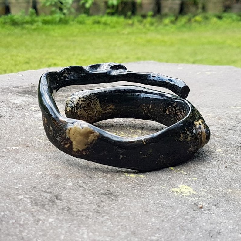 May include: A handcrafted, dark-stained wooden bracelet with a unique, organic shape. The bracelet features a swirling design with visible wood grain and lighter-colored patches, creating a natural, earthy aesthetic. The bracelet is a statement piece.