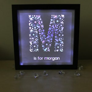 May include: A black framed light-up box with a white background. The letter "M" is cut out of the background and filled with a holographic pattern of stars, planets, and rockets. The text "is for morgan" is printed below the letter.