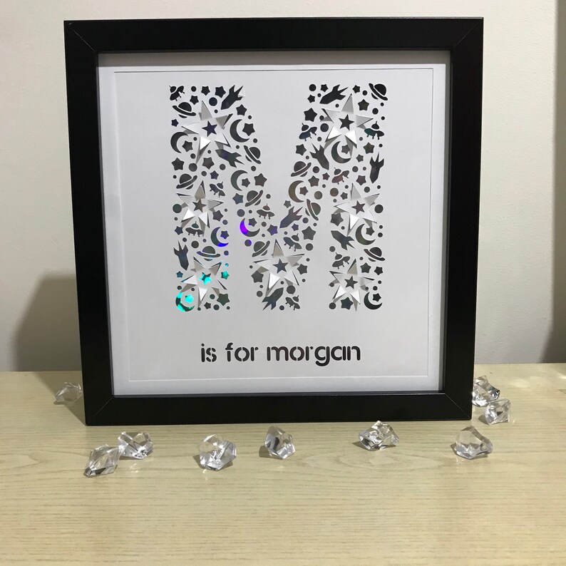 May include: A black framed picture with a white background. The letter "M" is cut out of the paper and filled with stars, moons, and rockets. The text "is for morgan" is below the letter.