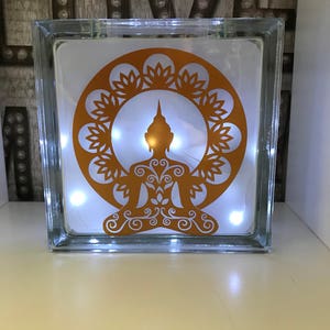 K&ouml;nnte beinhalten: Ein klarer Glasblock mit einem goldenen Buddha-Design und Lichterketten im Inneren. Der Buddha sitzt in der Lotusposition, umgeben von einem floralen Muster. Der Block ist beleuchtet und erzeugt ein warmes Leuchten.
