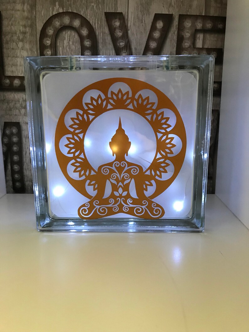 K&ouml;nnte beinhalten: Ein klarer Glasblock mit einem goldenen Buddha-Design und Lichterketten im Inneren. Der Buddha sitzt in der Lotusposition, umgeben von einem floralen Muster. Der Block ist beleuchtet und erzeugt ein warmes Leuchten.