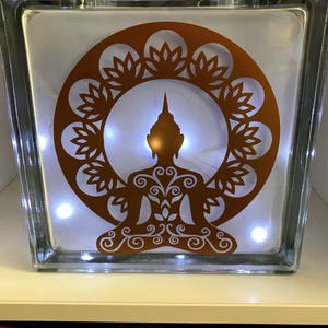 K&ouml;nnte beinhalten: Ein klarer Glasblock mit einem kupferfarbenen Buddha-Silhouette-Design. Der Buddha sitzt in meditativer Pose, umgeben von einem dekorativen Kreis. Kleine wei&szlig;e Lichter beleuchten das Innere und erzeugen ein warmes Leuchten. Ein dekorativer Artikel.