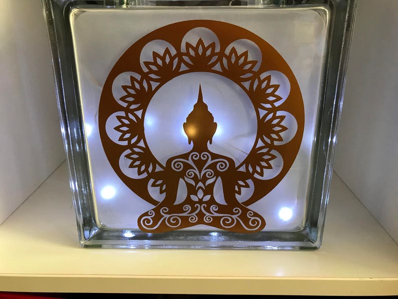 K&ouml;nnte beinhalten: Ein klarer Glasblock mit einem kupferfarbenen Buddha-Silhouette-Design. Der Buddha sitzt in meditativer Pose, umgeben von einem dekorativen Kreis. Kleine wei&szlig;e Lichter beleuchten das Innere und erzeugen ein warmes Leuchten. Ein dekorativer Artikel.