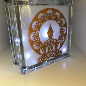 K&ouml;nnte beinhalten: Ein klarer Glasblock mit einem goldenen Buddha-Design und einer Lichterkette mit wei&szlig;en Lichtern im Inneren. Der Buddha sitzt in meditativer Pose, umgeben von einem floralen Muster. Ein dekoratives Element.