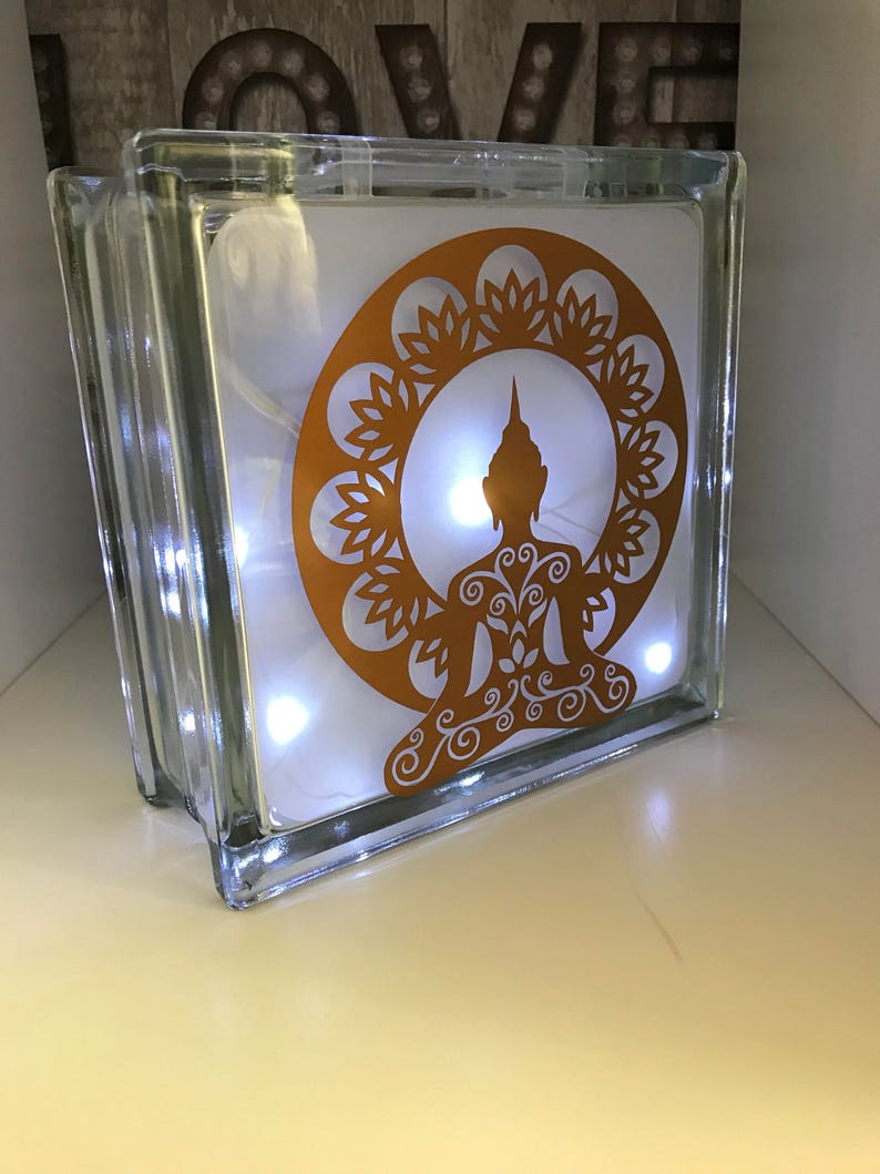 K&ouml;nnte beinhalten: Ein klarer Glasblock mit einem goldenen Buddha-Design und einer Lichterkette mit wei&szlig;en Lichtern im Inneren. Der Buddha sitzt in meditativer Pose, umgeben von einem floralen Muster. Ein dekoratives Element.