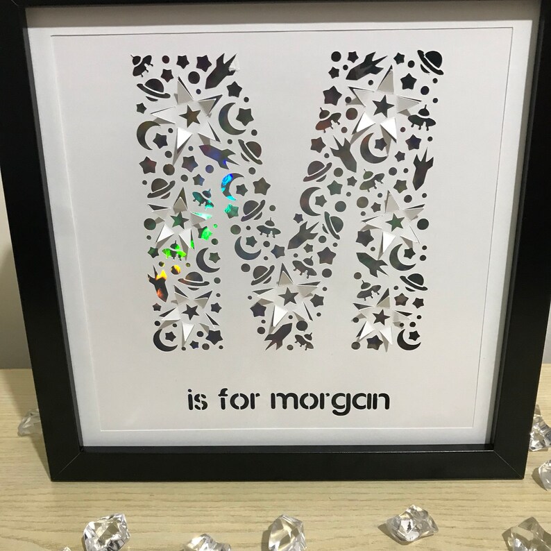 May include: A white framed picture with a black mat. The letter M is cut out of the white paper and filled with a holographic iridescent material. The letter M is surrounded by small cutouts of stars, moons, and planets. The text "is for morgan" is printed below the letter M.