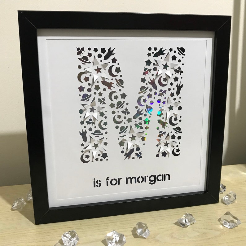 May include: A black framed picture with a white background. The letter M is cut out of the background and filled with a holographic iridescent material. The letter M is made up of small stars, moons, and planets. The text "is for Morgan" is below the letter M.