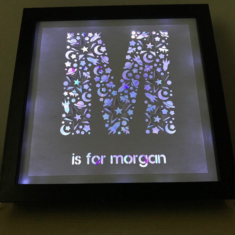 May include: A framed light-up letter M with a white background and a black frame. The letter M is made of iridescent paper cutouts of stars, moons, rockets, and planets. The text "is for morgan" is below the letter.