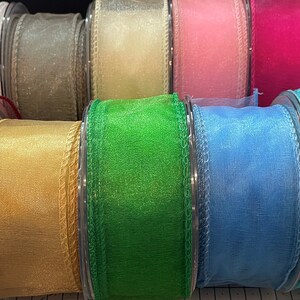 K&ouml;nnte beinhalten: Eine Sammlung farbenfroher, durchsichtiger Bandspulen. Die B&auml;nder sind in verschiedenen Farben erh&auml;ltlich, darunter Schwarz, Wei&szlig;, Gold, Rosa und Blau. Das Band wird wahrscheinlich f&uuml;r Bastelarbeiten, Geschenkverpackungen oder Dekorationen verwendet.