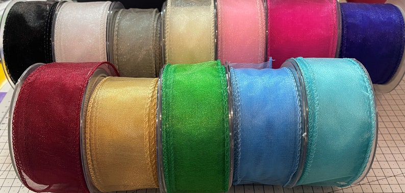 K&ouml;nnte beinhalten: Eine Sammlung farbenfroher, durchsichtiger Bandspulen. Die B&auml;nder sind in verschiedenen Farben erh&auml;ltlich, darunter Schwarz, Wei&szlig;, Gold, Rosa und Blau. Das Band wird wahrscheinlich f&uuml;r Bastelarbeiten, Geschenkverpackungen oder Dekorationen verwendet.