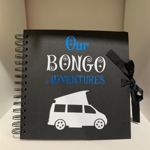 May include: Black scrapbook with a white silhouette of a camper van and the text "Our Bongo Adventures" in white with blue glitter accents. The scrapbook has a black ribbon tied around it.