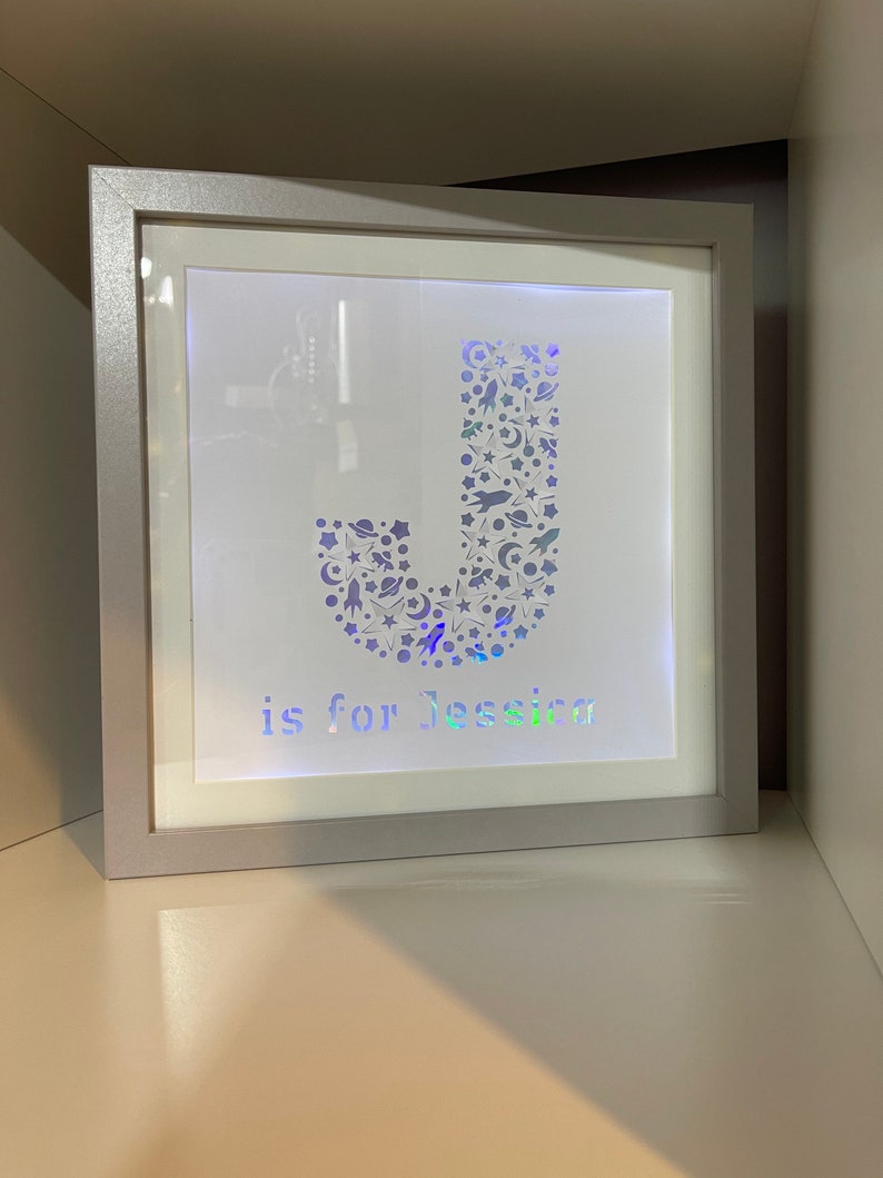 May include: A silver framed light-up letter J with a space-themed design. The letter is made up of stars, planets, and rockets. The text "J is for Jessica" is below the letter.