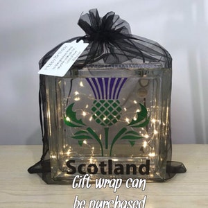 K&ouml;nnte beinhalten: Ein dekorativer Glasblock mit einem Disteldesign in Gr&uuml;n und Lila, darunter das Wort "Scotland". Der Block ist mit schwarzem T&uuml;ll umwickelt und enth&auml;lt Lichterketten. Ein Etikett lautet "Happy Birthday".