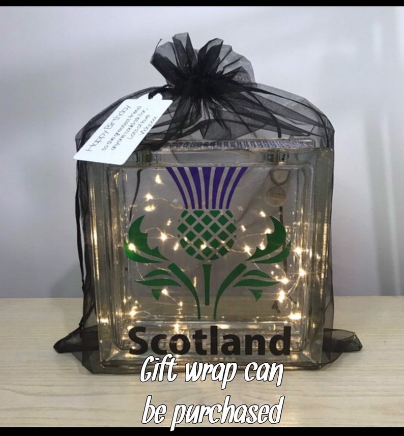 K&ouml;nnte beinhalten: Ein dekorativer Glasblock mit einem Disteldesign in Gr&uuml;n und Lila, darunter das Wort "Scotland". Der Block ist mit schwarzem T&uuml;ll umwickelt und enth&auml;lt Lichterketten. Ein Etikett lautet "Happy Birthday".