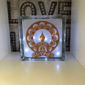 K&ouml;nnte beinhalten: Ein klarer Glasblock mit einem goldfarbenen Buddha-Design und einer Lichterkette mit wei&szlig;en Lichtern im Inneren. Der Buddha befindet sich in einem kreisf&ouml;rmigen Blumendesign. Im Hintergrund steht das Wort "LOVE" in gro&szlig;en, beleuchteten Buchstaben.