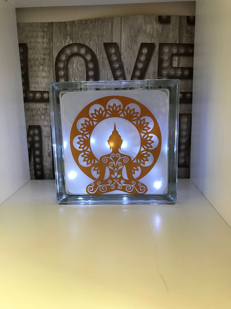 K&ouml;nnte beinhalten: Ein klarer Glasblock mit einem goldfarbenen Buddha-Design und einer Lichterkette mit wei&szlig;en Lichtern im Inneren. Der Buddha befindet sich in einem kreisf&ouml;rmigen Blumendesign. Im Hintergrund steht das Wort "LOVE" in gro&szlig;en, beleuchteten Buchstaben.