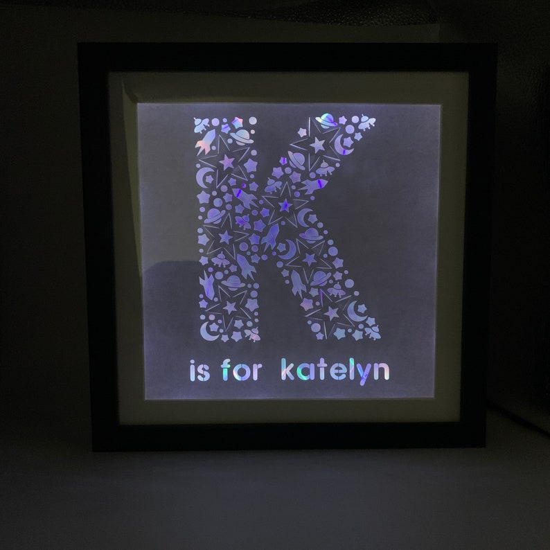 May include: A black framed light-up sign with a white letter K made of iridescent stars, moons, and rockets. The text "is for katelyn" is below the letter.