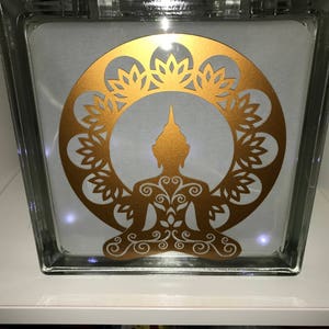 K&ouml;nnte beinhalten: Ein klarer Glasblock mit einem goldenen Buddha-Silhouette-Design. Der Buddha sitzt in meditativer Pose, umgeben von einem floralen Kranz. Das Design wird von kleinen Lichtern im Inneren des Blocks beleuchtet.