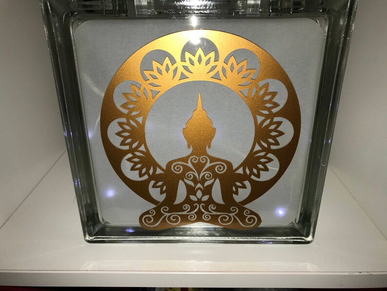 K&ouml;nnte beinhalten: Ein klarer Glasblock mit einem goldenen Buddha-Silhouette-Design. Der Buddha sitzt in meditativer Pose, umgeben von einem floralen Kranz. Das Design wird von kleinen Lichtern im Inneren des Blocks beleuchtet.