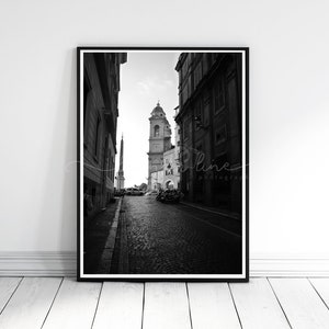May include: Black and white framed photograph of a cobblestone street in Rome, Italy. The image captures a perspective view of buildings, a church with a bell tower, and an obelisk. The photograph is mounted in a black frame and leans against a white wall.