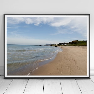 Può includere: Una stampa incorniciata di una scena di spiaggia. L'immagine mostra una spiaggia sabbiosa con dolci onde, un cielo azzurro e una costa lontana. La stampa è in una cornice nera ed è esposta contro un muro bianco e un pavimento in legno.
