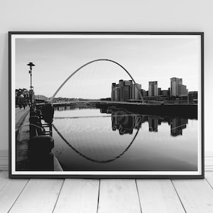 Può includere: Una fotografia in bianco e nero di un ponte ad arco moderno su un fiume in una città. Il ponte si riflette nell'acqua. Lo skyline della città è visibile sullo sfondo.