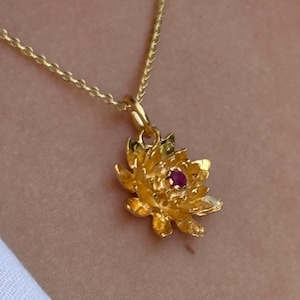 May include: A gold-colored necklace with a flower pendant. The pendant features a detailed floral design with a small, red gemstone at its center. The necklace is worn against a white fabric.