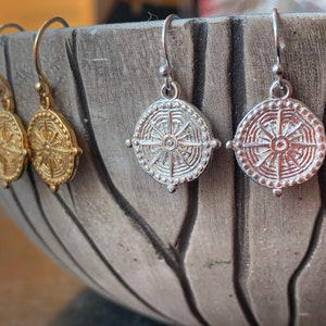 May include: A pair of gold and silver earrings with a circular design featuring a sunburst pattern. The earrings are hanging from a bowl.