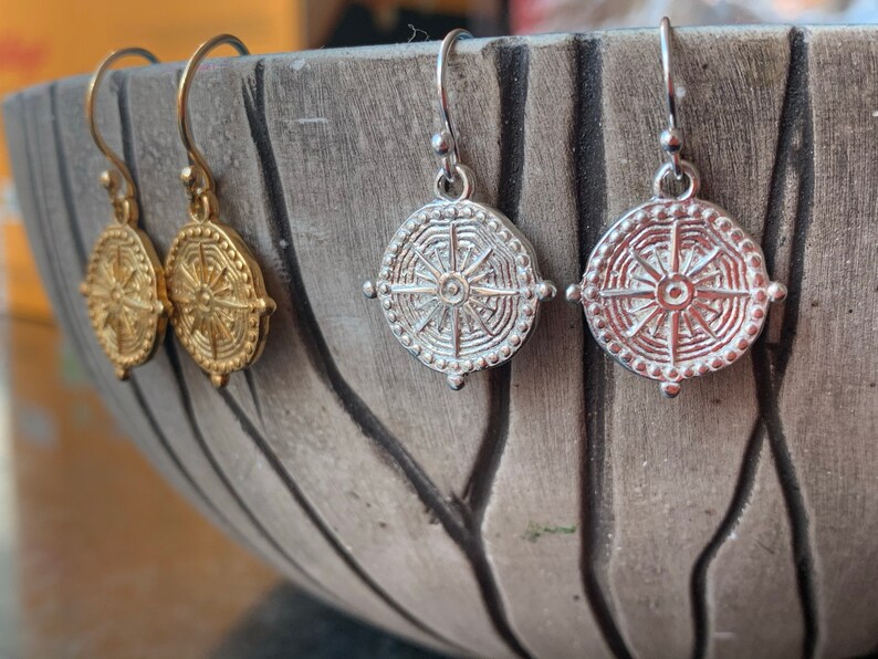 May include: A pair of gold and silver earrings with a circular design featuring a sunburst pattern. The earrings are hanging from a bowl.