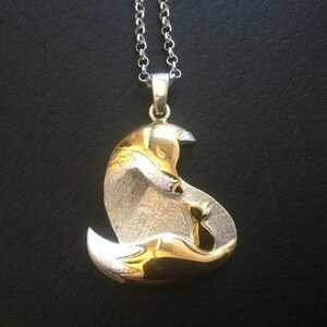 May include: Silver and gold-toned pendant necklace featuring a stylized fox design. The fox is depicted in a curled position, with intricate detailing. The pendant hangs from a delicate silver chain, creating an elegant accessory.