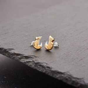 May include: A pair of gold-colored fox stud earrings with silver posts. The earrings have a two-tone design, with the fox's body in a shiny gold and the lower portion in silver. The earrings are displayed on a dark gray surface.