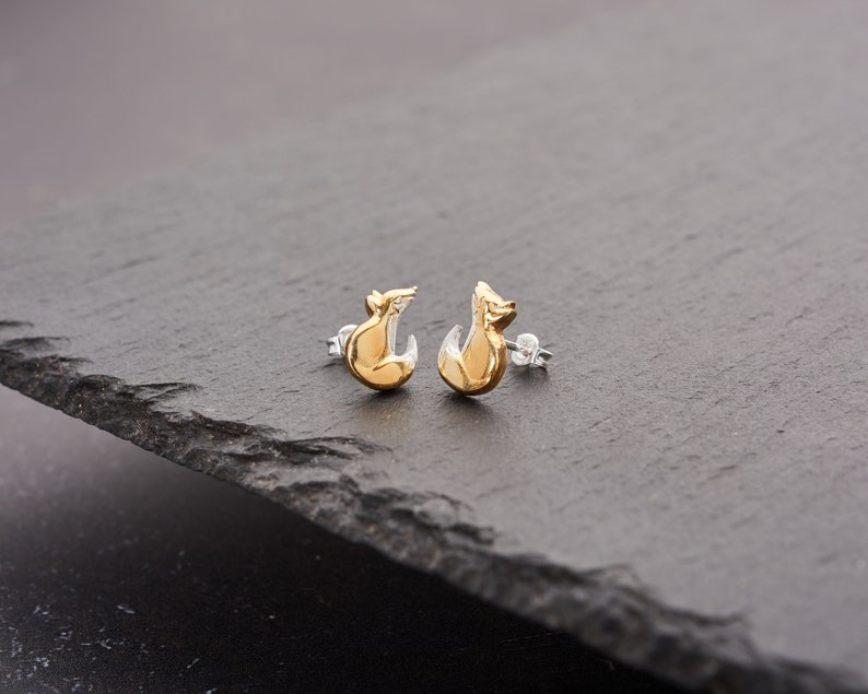 May include: A pair of gold-colored fox stud earrings with silver posts. The earrings have a two-tone design, with the fox's body in a shiny gold and the lower portion in silver. The earrings are displayed on a dark gray surface.