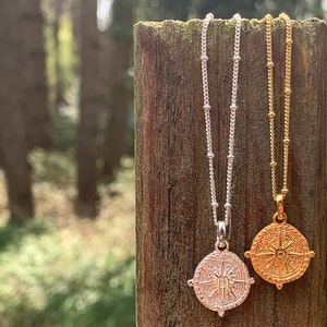 May include: Two silver and gold necklaces with compass pendants hanging on a wooden post. The pendants feature a detailed compass design with a sunburst in the centre.
