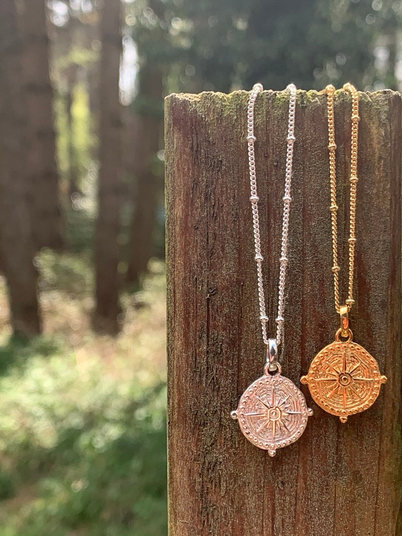 May include: Two silver and gold necklaces with compass pendants hanging on a wooden post. The pendants feature a detailed compass design with a sunburst in the centre.