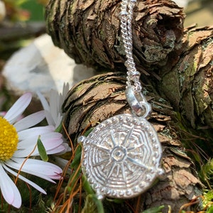 May include: A silver pendant necklace with a detailed circular design featuring a sunburst pattern. The necklace is on a chain with small beads and is resting on a bed of moss and pine cones.
