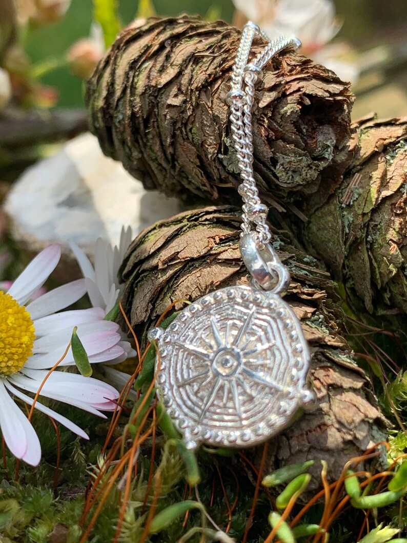 May include: A silver pendant necklace with a detailed circular design featuring a sunburst pattern. The necklace is on a chain with small beads and is resting on a bed of moss and pine cones.
