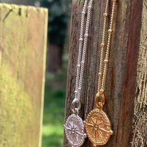 May include: Two necklaces with compass pendants. The necklace on the left has a silver chain and a silver compass pendant. The necklace on the right has a gold chain and a gold compass pendant.