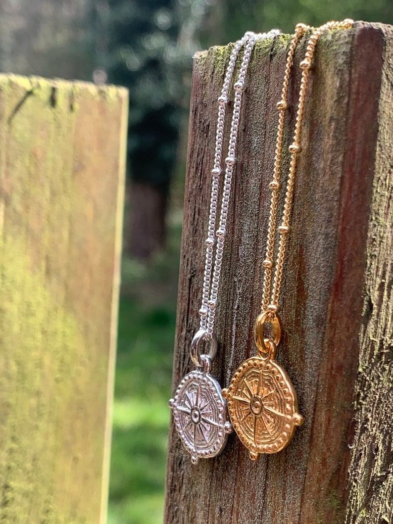 May include: Two necklaces with compass pendants. The necklace on the left has a silver chain and a silver compass pendant. The necklace on the right has a gold chain and a gold compass pendant.