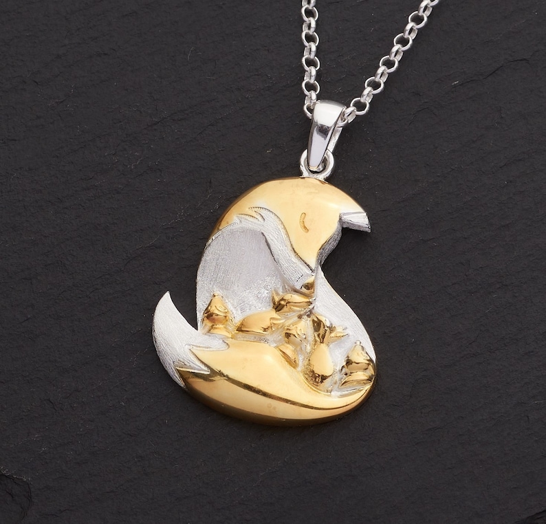 May include: A silver and gold-tone pendant necklace featuring a fox and its kits. The pendant has a brushed silver finish with gold-tone accents. The fox and kits are nestled together in a crescent shape, suspended from a silver chain.