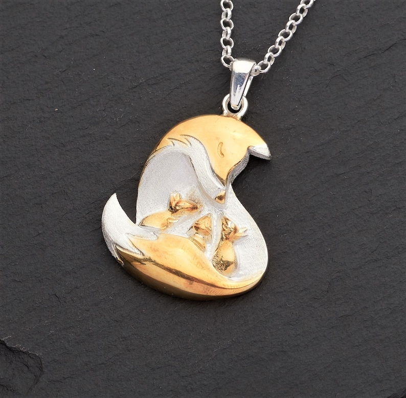 May include: A silver necklace with a pendant featuring a gold and silver fox design. The pendant showcases a mother fox with three smaller foxes nestled within. The necklace is displayed on a dark, textured surface.