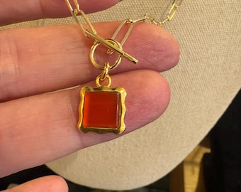 Pode incluir: Colar dourado com um pingente quadrado laranja em uma moldura decorativa. O colar tem um fecho de alternância e uma corrente com elos retangulares. O pingente é preso por um anel dourado.