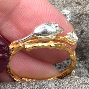 May include: A gold-coloured ring with a branch-like band and a silver-coloured top. The ring features a small, silver-coloured bud and a textured design, creating a nature-inspired aesthetic.