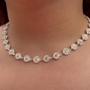 May include: A silver necklace featuring a daisy flower design. Each daisy has white petals and a yellow center. The necklace is worn around the neck and is a delicate piece of jewelry.